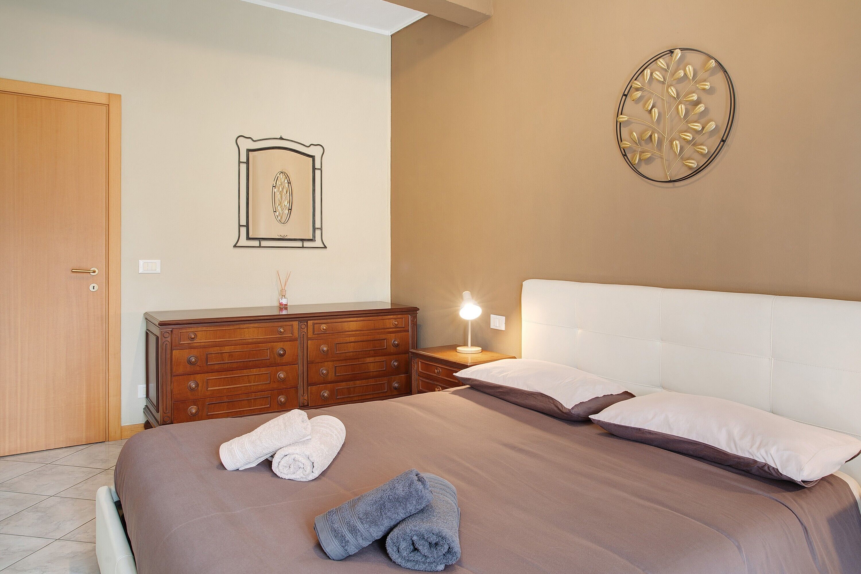 Una camera da letto con un letto, una cassettiera, un comodino con una lampada, un quadro appeso al muro e una porta.