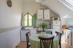 Een compacte keuken met een eettafel, stoelen, een potplant en een raam met groene luiken.
