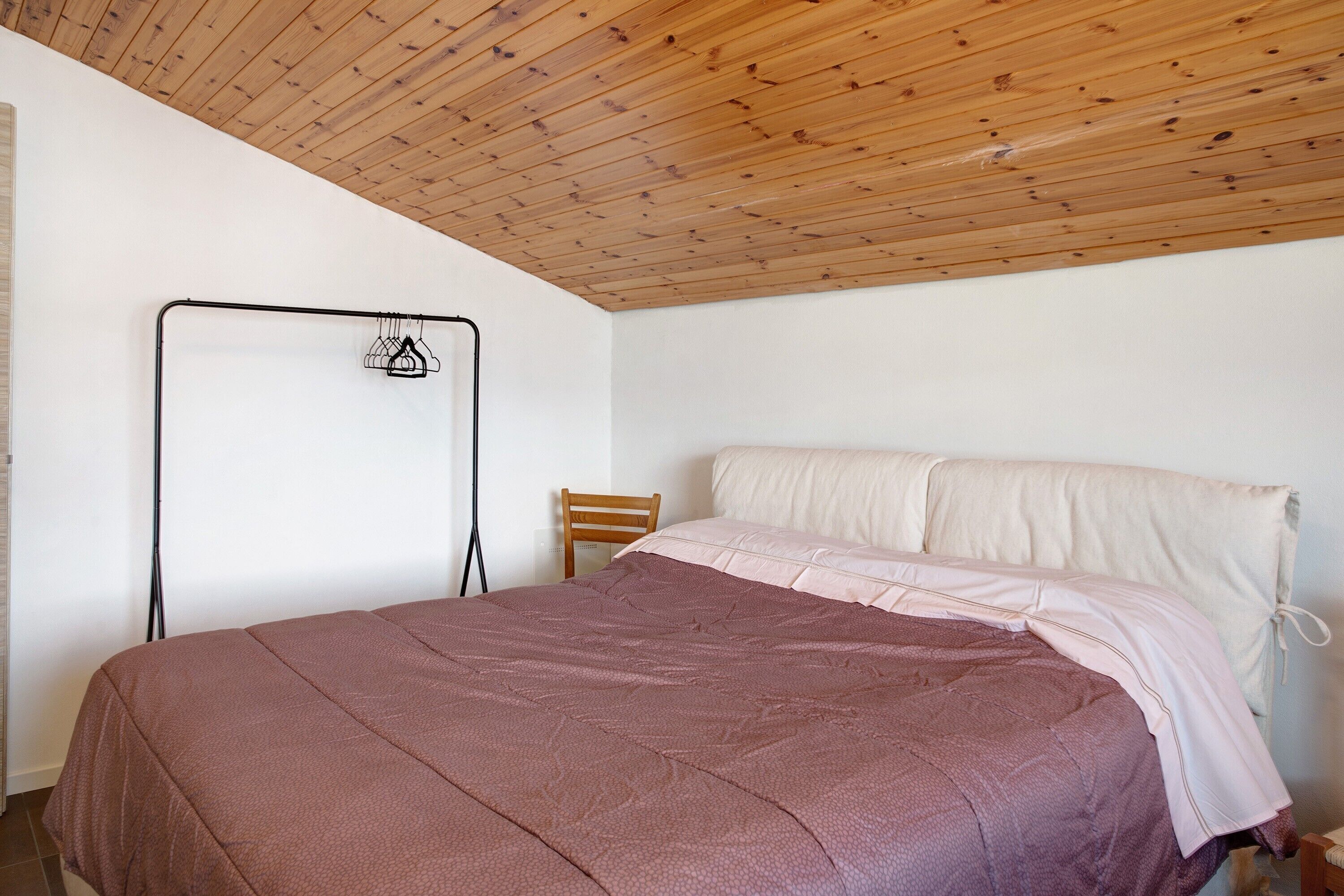 Un dormitorio con techo de madera, cama con colcha rosa y marrón, una silla y un tendedero de metal.