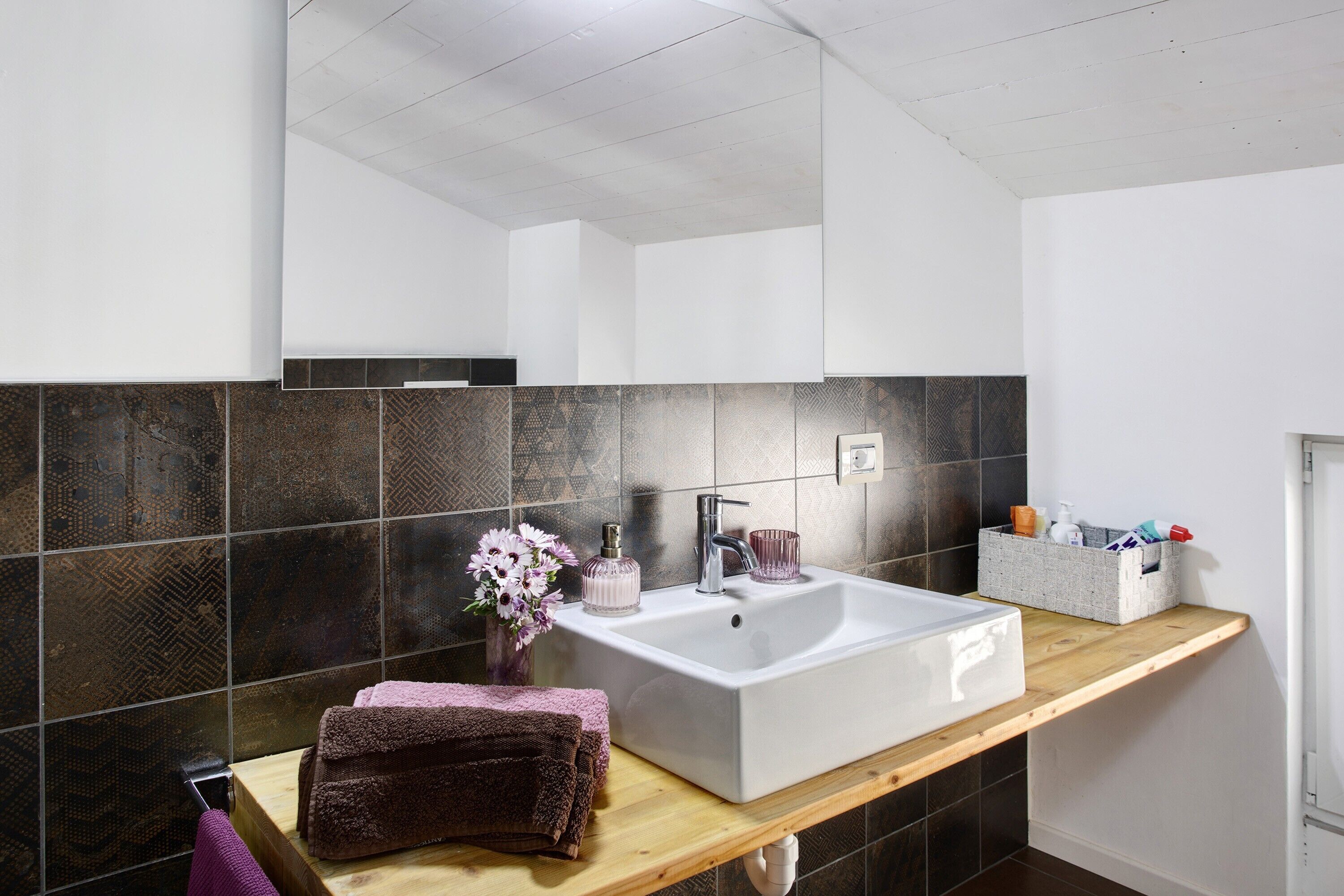 Un baño con un lavabo grande de color blanco, baldosas oscuras con diseños, una encimera de madera y un espejo sobre el lavabo.