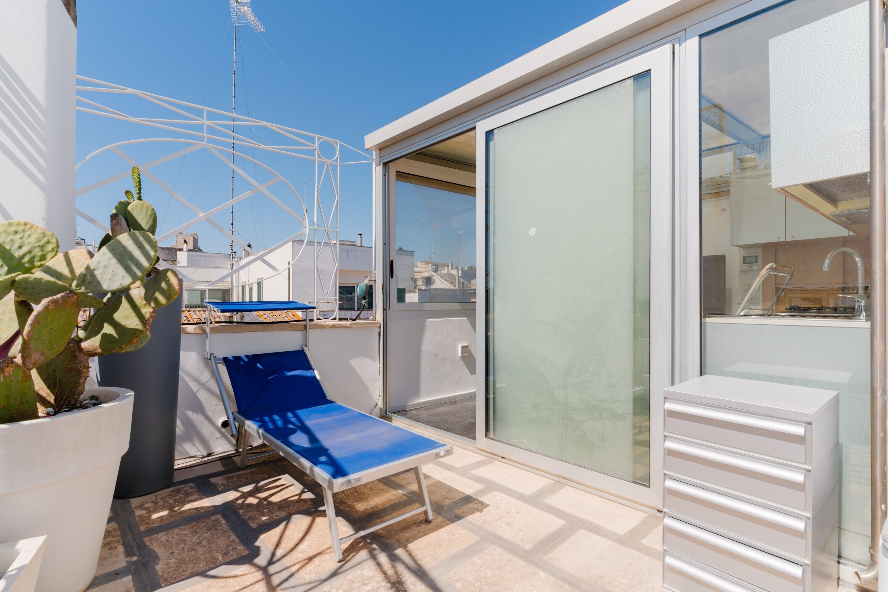 A balcony with a blue lounge chair, a potted cactus, and a sliding glass door leading to a kitchen area.