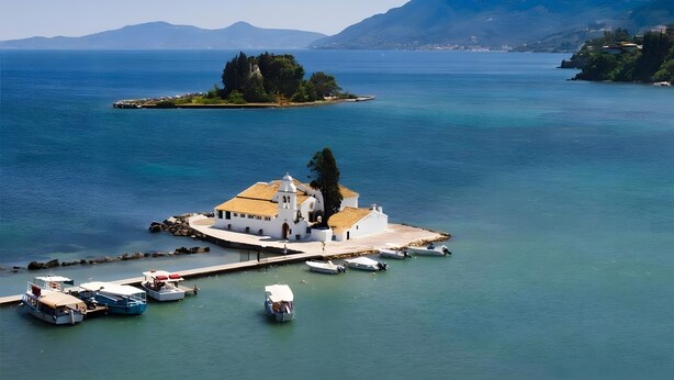 A coastal area with a white building with a red roof, boats docked at a pier, and a small island with trees.