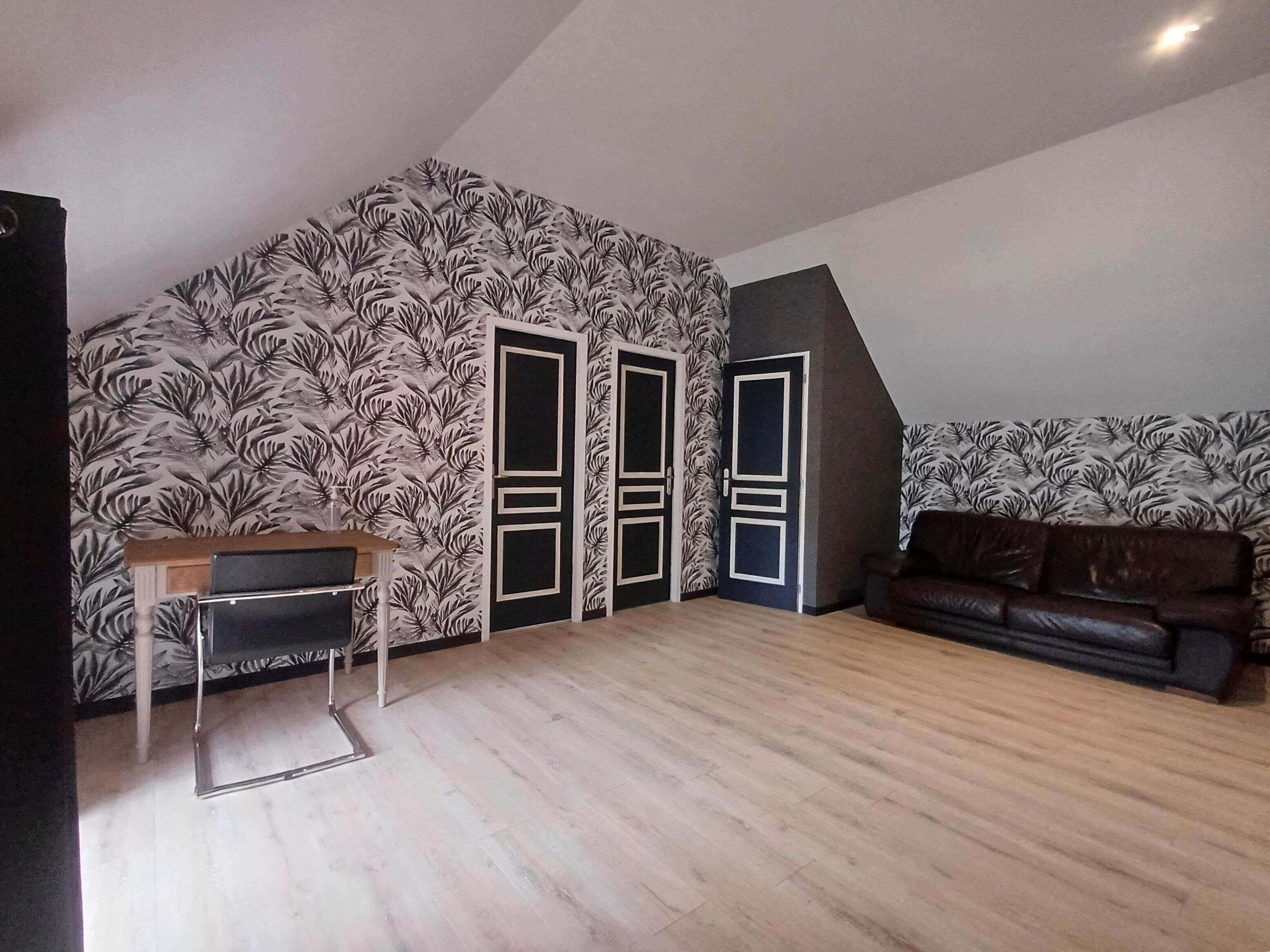 Ein Zimmer mit schrägen Wänden, Holzboden, einer schwarz-weißen Wandverkleidung, einem braunen Ledersofa und einem kleinen Tisch mit Stuhl.