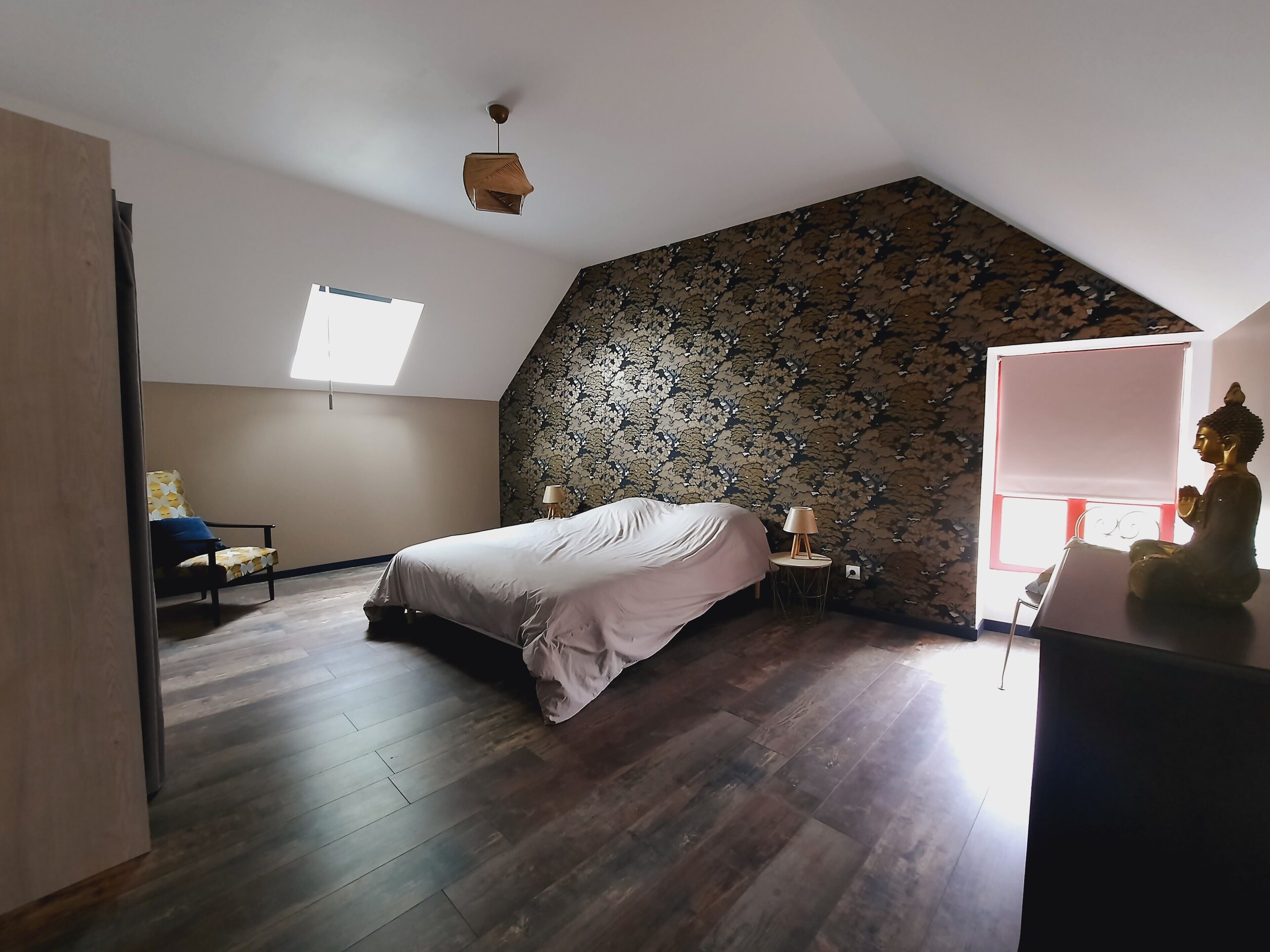 Ein Schlafzimmer mit schräg stehender Decke, einem Bett, einem Sessel, einem kleinen Tisch mit Lampe und einer Statue.