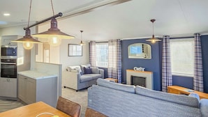 A modern kitchen with built-in appliances, a dining area with a fruit bowl, a living room with a sofa and fireplace, and a bedroom with a bed and curtains. - Bute Lodge with Hot Tub (Lendalfoot)