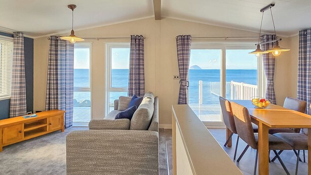 A room with a sofa, a wooden table, and a bench. There are curtains and a view of the sea. - Bute Lodge with Hot Tub (Lendalfoot)