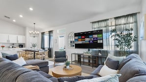 Ein modernes Wohnzimmer mit einem Flachbildfernseher, grauen Sofas, einem Couchtisch und einem Essbereich mit einer Küche im Hintergrund.