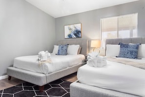 A hotel room with two beds, a gray sofa, a black and white rug, a painting on the wall, and a window with blinds.