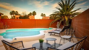 A swimming pool with lounge chairs and a small table, surrounded by palm trees and a brick wall.