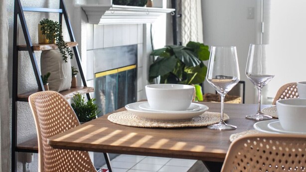 A dining area with a wooden table, white plates, and wine glasses. There is a wicker chair and a shelf with plants.