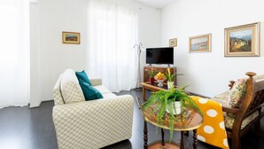 Een woonkamer met een bank, een bijzettafeltje met planten, een televisie en ingelijste foto's aan de muur.