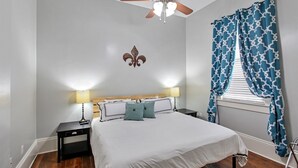 A bedroom with a bed, two bedside tables, a ceiling fan, and a window with curtains. - The Tchoup Shop (New Orleans)