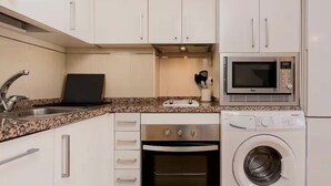 Une cuisine compacte avec des meubles blancs, un plan de travail en granit, un four encastré, un four à micro-ondes et un lave-linge à chargement frontal.