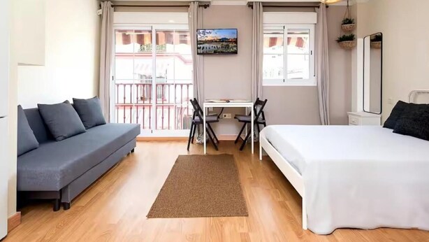 Une chambre d’hôtel moderne avec un canapé gris, un lit, un bureau avec une chaise et un balcon offrant une vue.