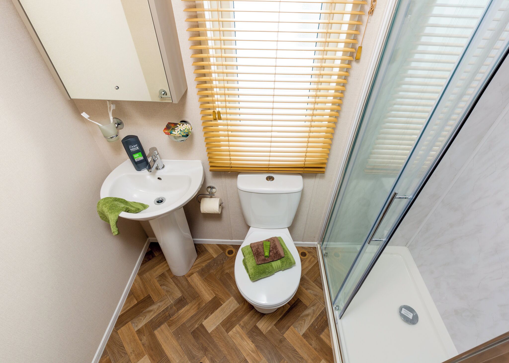 Une salle de bain avec un plancher en bois, un lavabo blanc, des toilettes et une cabine de douche en verre.