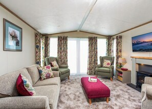 A cozy living room with a sofa, two armchairs, a red ottoman, a fireplace, a TV, and floral curtains. - 2 bedroom accommodation in Nr Kintore (Nr Kintore)