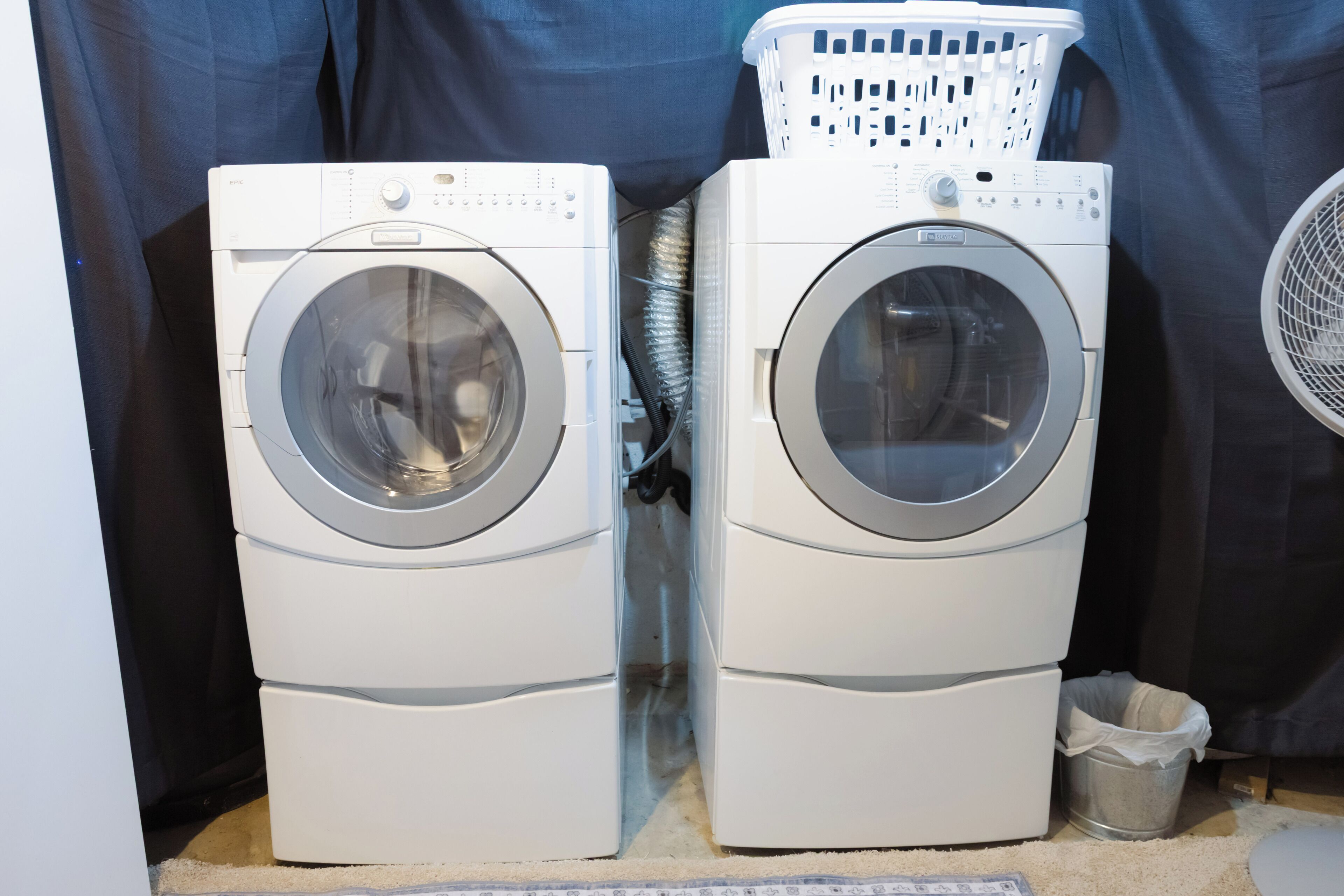 Deux machines à laver à chargement frontal, superposées, avec un panier blanc sur le dessus, une poubelle en métal et un ventilateur sur la droite.