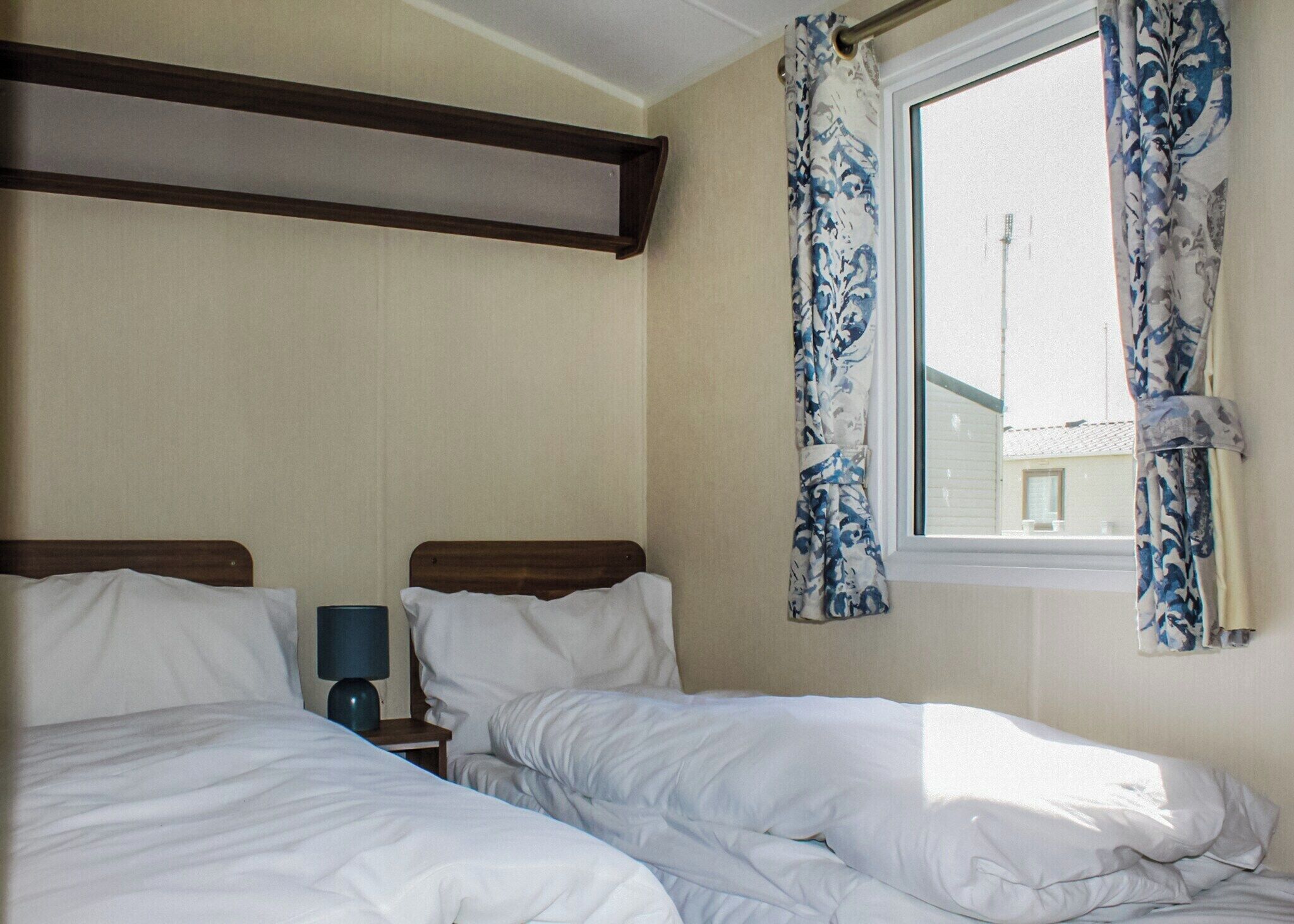 A bedroom with two beds, a window with curtains, a nightstand with a lamp, and a wooden beam above the beds.