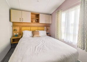 A hotel room with a bed, wooden headboard, bedside tables, a nightstand, a shelf with a heart-shaped decoration, and a window with patterned curtains.