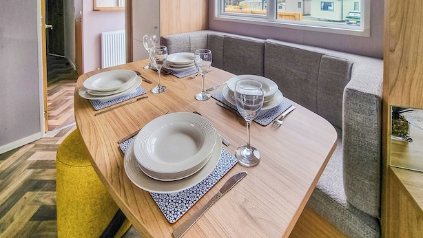 Une table à manger dressée avec des assiettes, des verres et des couverts, une fenêtre donnant sur des maisons et une porte menant à une autre pièce.