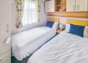 A room with two beds, a window with patterned curtains, a bedside table with a lamp, and wooden cabinets above the beds.