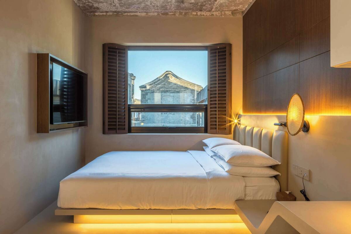 Una cama bien tendida en una habitación con un amplio ventanal, un televisor montado en la pared y una lámpara de pared original.