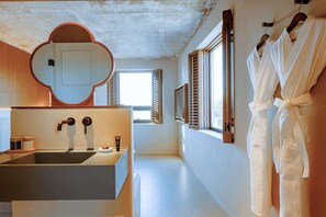 Ein modernes Badezimmer mit einem großen Waschbecken, Holzjalousien und weißen Bademänteln, die an der Wand hängen.