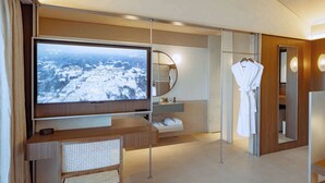 Une chambre d’hôtel moderne dotée d’un grand téléviseur à écran plat, d’une coiffeuse en bois, d’un miroir et d’un peignoir accroché à la porte.