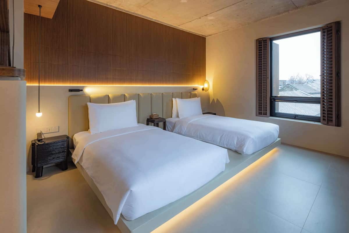 Una habitación de hotel moderna con dos camas, techo de madera, un ventanal con contraventanas y una mesita de noche con lámpara.
