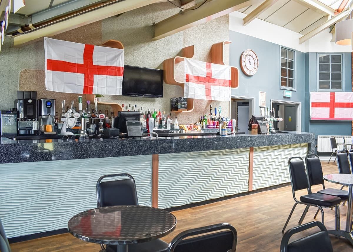 A bar area with a counter, bar stools, and flags. There is a clock on the wall and a television mounted above the counter.