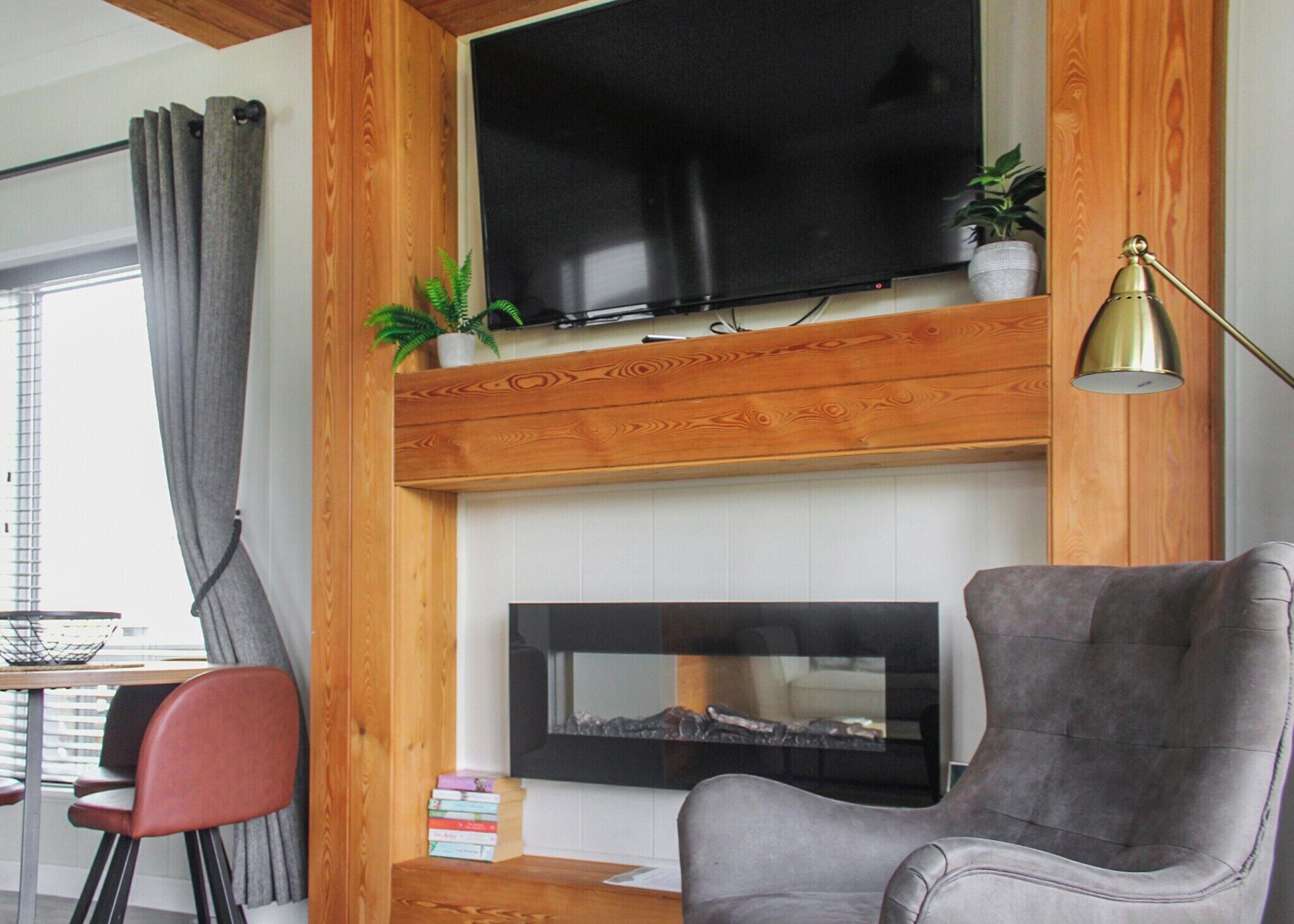 A living room with a flat-screen TV mounted on a wooden wall, a fireplace below, a grey armchair, a small table with books, and a floor lamp.