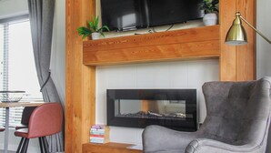 Een woonkamer met een flatscreen televisie aan een houten wand, een haard eronder, een grijze fauteuil, een klein tafeltje met boeken en een vloerlamp.