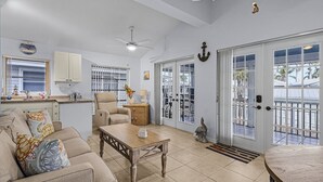 A spacious living area with a sofa, coffee table, and a dining table. There is a kitchen area with cabinets and a window. The room has a ceiling fan and a view of the outside through large glass doors.