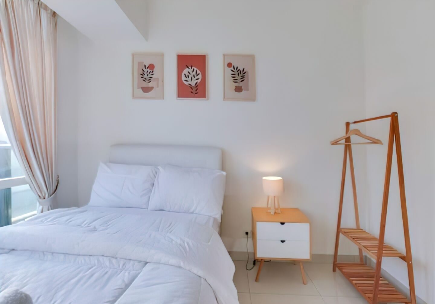 A bedroom with a bed, a bedside table with a lamp, a wooden hanger shelf, and three framed artworks on the wall.