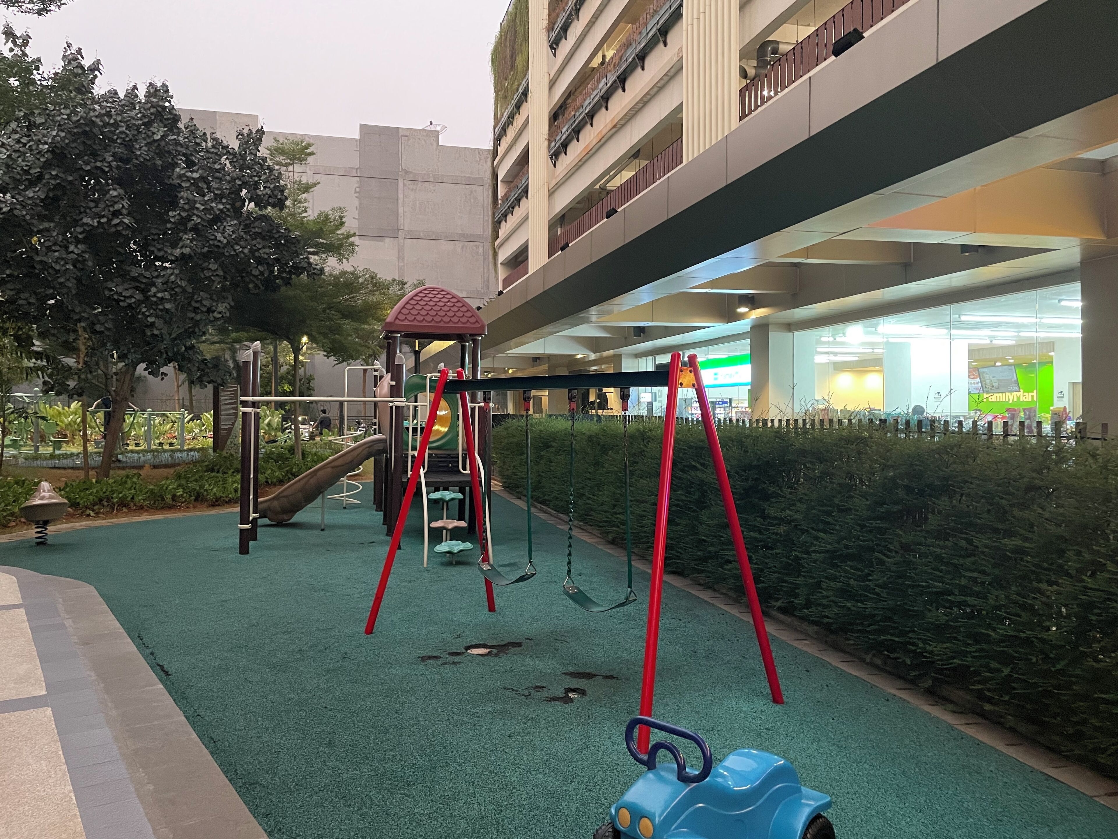 A playground with swings and a slide, surrounded by greenery and buildings in the background.