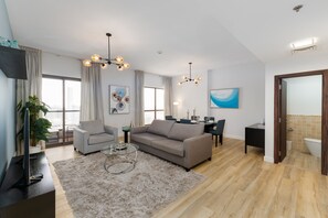 A modern living room with a grey sofa, a coffee table, and a TV on a black stand. There is a dining area with a table and chairs, and a kitchen area with a sink and countertop.