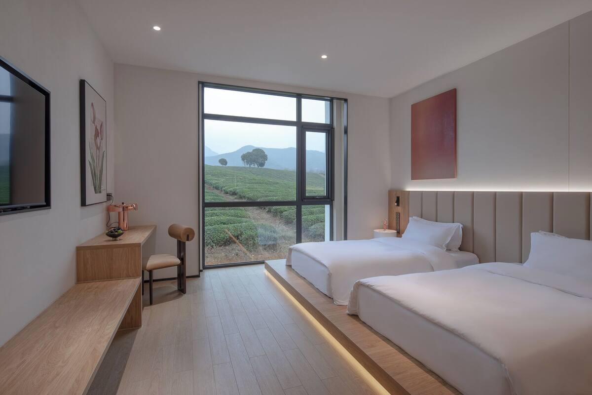 A modern hotel room with two beds, a desk, and a large window offering a view of a green landscape.