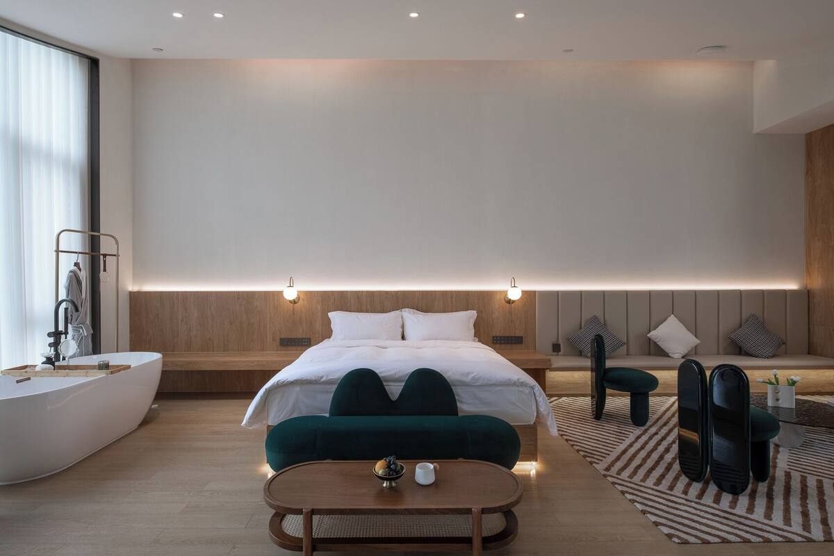 A modern hotel room with a large bed, a wooden headboard, a built-in bathtub, and a seating area with a table and chairs.