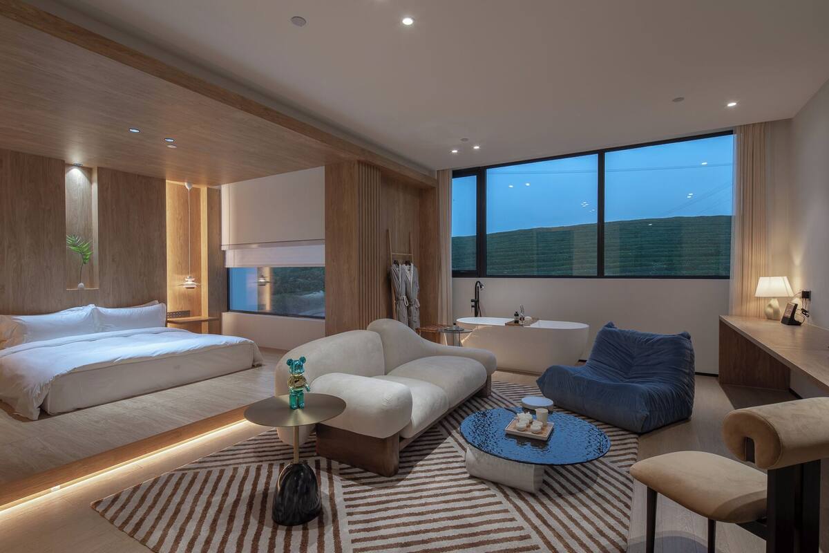 A modern hotel room with a large bed, a sofa, a blue armchair, a round coffee table, and a bathtub.