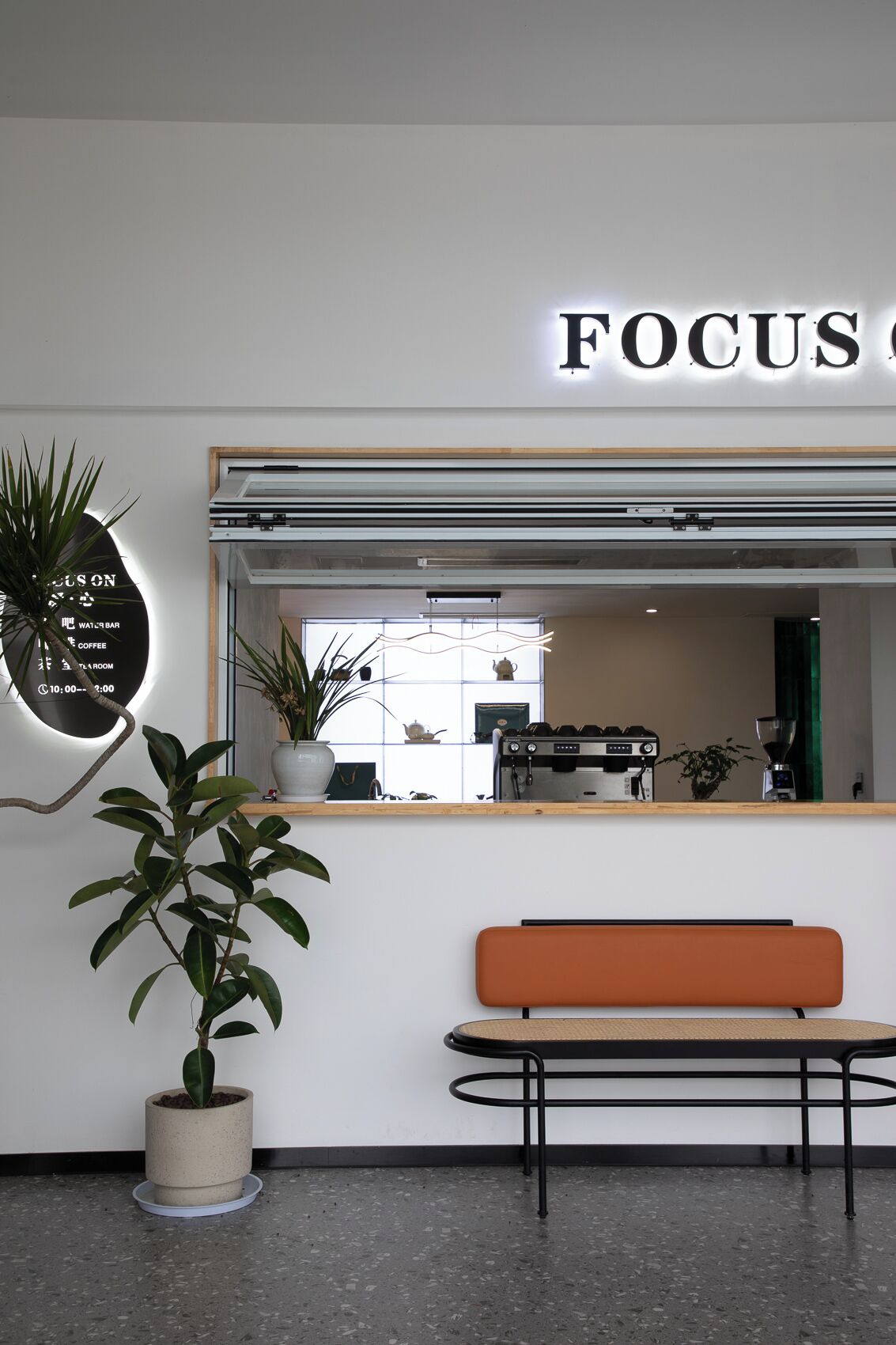 Ein modernes Interieur mit einem 'FOCUS'-Schild, einer Bank, einem Blumentopf und einer Kaffeemaschine, die durch ein Fenster sichtbar ist.