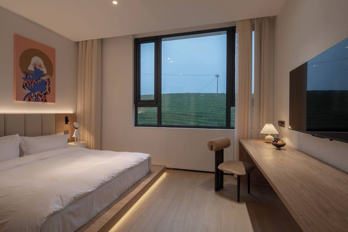 A hotel room with a large bed, a desk with a chair, a TV, and a window with a view of a green field.