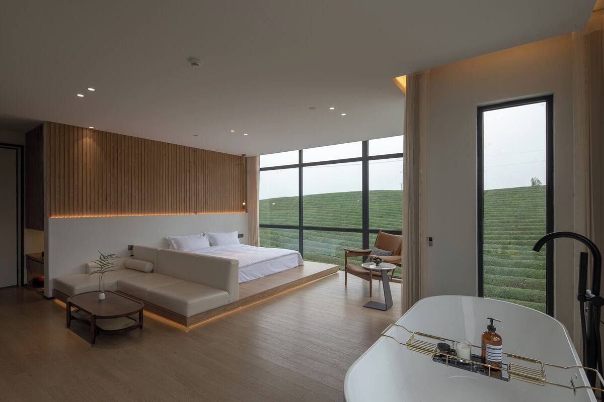 A modern bedroom with a large bed, a sofa, a small table, and a view of a green landscape through large windows.