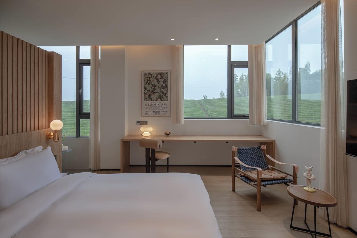 A modern hotel room with a large bed, a desk, and a chair. There is a window with a view of a green landscape.