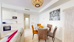 Une cuisine moderne avec des meubles blancs, un four à micro-ondes et un four traditionnel. On y trouve un coin repas avec une table ronde, des chaises et un saladier rempli d’oranges. Un grand tableau représentant un chat est accroché au mur.
