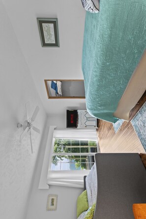 Une pièce dotée d’un ventilateur de plafond, d’une fenêtre donnant sur des arbres, d’un lit recouvert d’une courtepointe turquoise, d’un tableau encadré au mur et d’une petite table sur laquelle se trouve une horloge.
