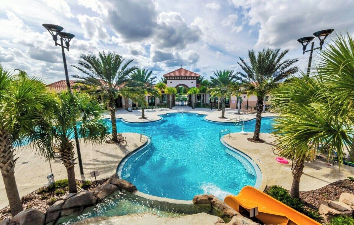 A large outdoor swimming pool surrounded by palm trees and a building with a red-tiled roof.