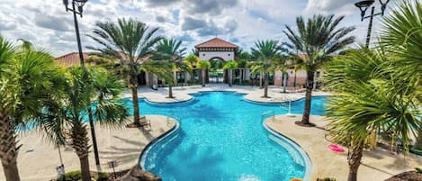 A large outdoor swimming pool surrounded by palm trees and a building with a red-tiled roof.