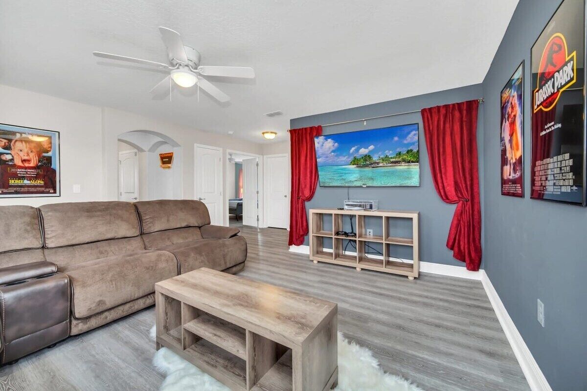 A living room with a brown sofa, a wooden coffee table, a TV on the wall, and red curtains.