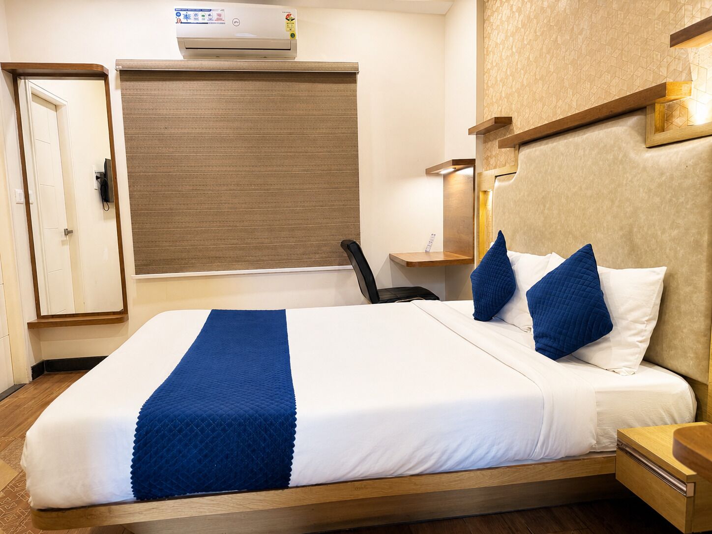 A hotel room with a bed, a desk, a chair, a wall-mounted air conditioner, and a window with a blind.