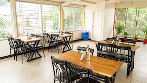 A dining area with multiple tables and chairs, large windows with blinds, and potted plants. - Hotel Rize Inn (Bengaluru)